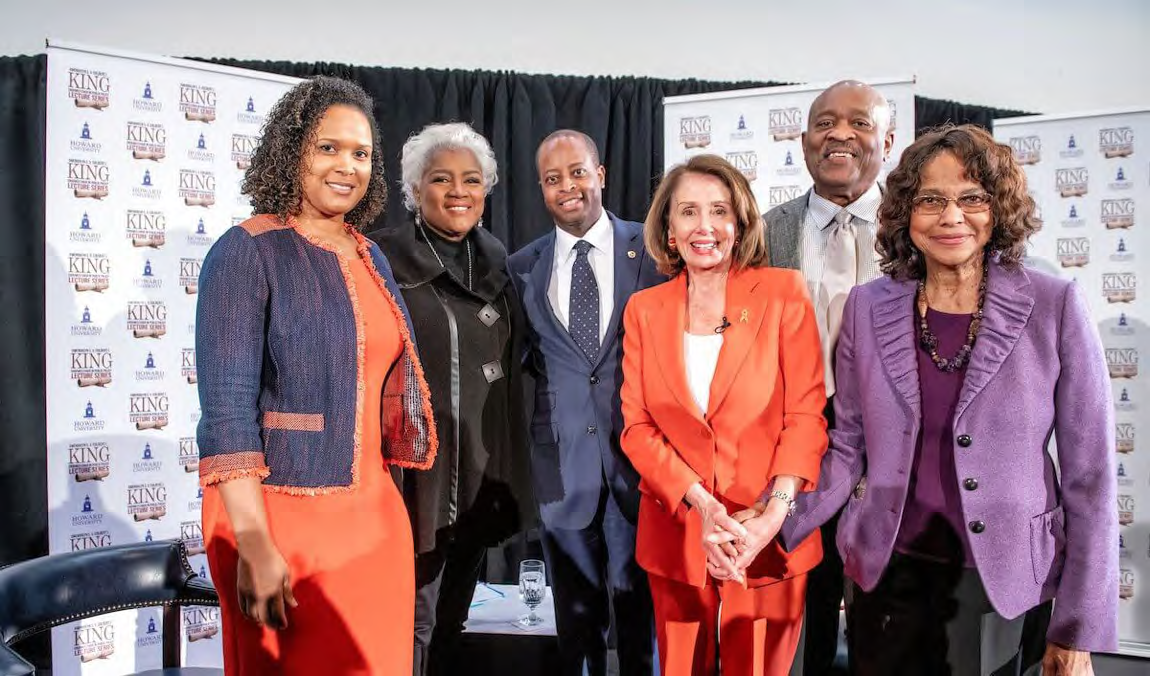 Group of smiling people at the King series event, including President Wayne A. I. Frederick standing at the center, United States Representative Nancy Pelosi in red with a white shirt, Gwendolyn S. King in purple standing beside her, and Colbert I. King standing behind them. The Gwendolyn S. and Colbert I. King Chair Endowed Chair in Public Policy Lecture Series logo is printed on the walls behind them.